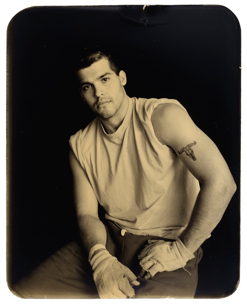 A black and white photograph of a young, light-skinned man sitting facing the camera with his shirt-sleeves rolled up to his shoulders.
