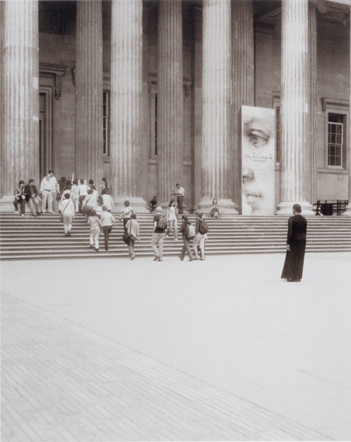 A black-and-white photograph shows a crowd of people walking up the steps of a large classical-style building while a figure in a long modest black shift stands watching the crowd.