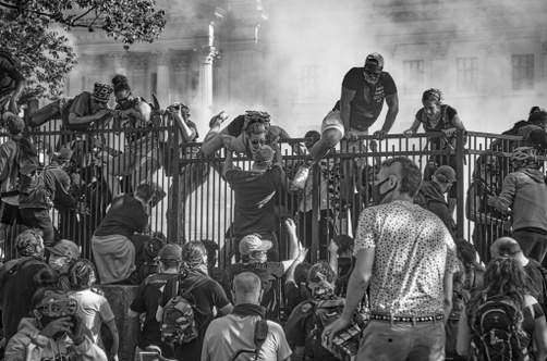 A black and white photo of protestors climbing over an iron fence in front of a colonial-stye building towards a group of protestors on the other side. A white fog curls up behind the protestors climbing the fence.