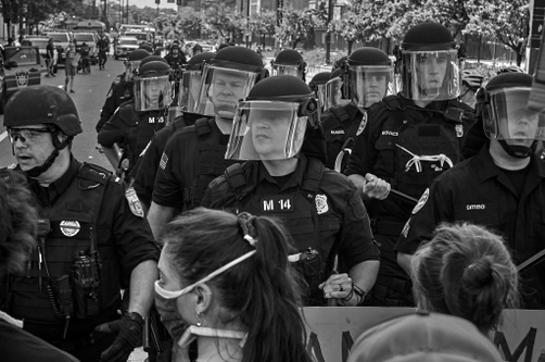 A black and white photograph of light-skinned policemen in black gear, wearing face shields, and carrying batons lined up facing a group of protestors.