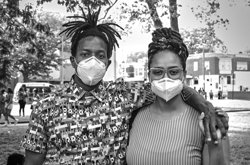A black and white photograph of two young, dark-skinned people, one male and one female, wearing white masks standing next to each other in front of an urban landscape.