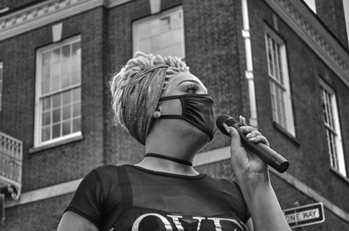 A young, tanned woman wearing a black mask speaks into a microphone in front of a brick building.