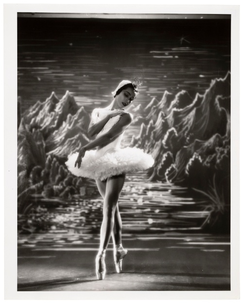 A black-and-white photograph of a ballet dancer in a tutu performing in front of an illustrated backdrop depicting a mountainous landscape.