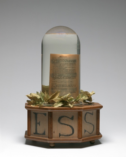 A book submerged in water inside a dome-shaped terrarium is situated on top of a laurel wreath and an octagonal, wooden base with letters C, E, S.