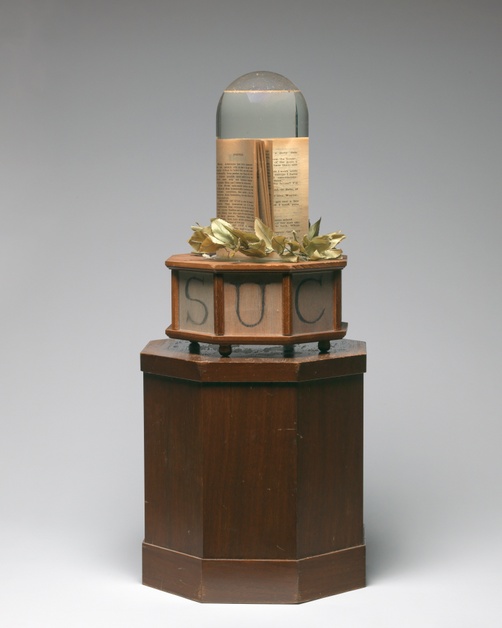 A book submerged in water inside a dome-shaped terrarium is situated on top of a laurel wreath and an octagonal, wooden base with letters C, E, S.