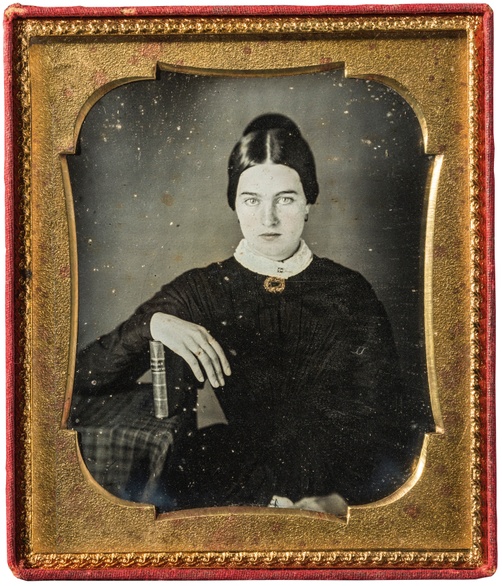 Black and white photographic portrait of an adult woman with her hand resting atop a book, all within a red and gold frame. 