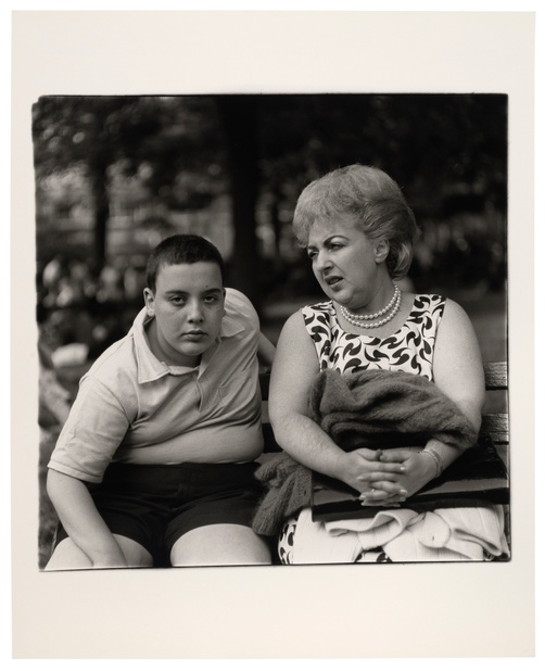 In a black and white photograph, a young boy intensely looks at us while sitting on a bench next to his mother who looks to the right.