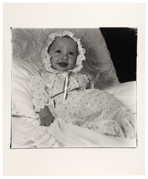 In a black and white photograph, a baby gazes upward with an open-mouthed smile and a bonnet tied underneath their chin while reclining on a seat covered in lace fabric.