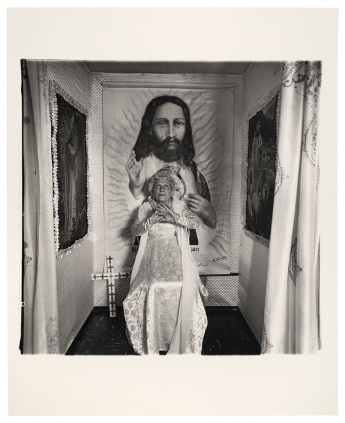 In a black and white photograph, a woman in a white dress looks up and crosses her hands in front of her chest in a small room featuring tapestries of Jesus.