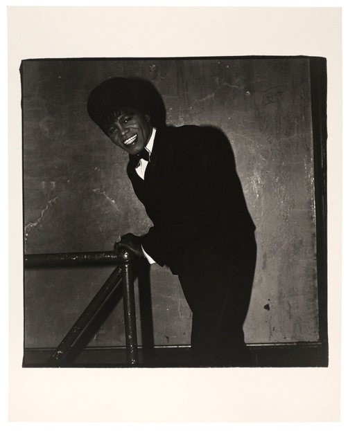 In a black and white photograph, a Black man leans against a railing while wearing a black tux with a wide and radiant smile in a dimly lit room.