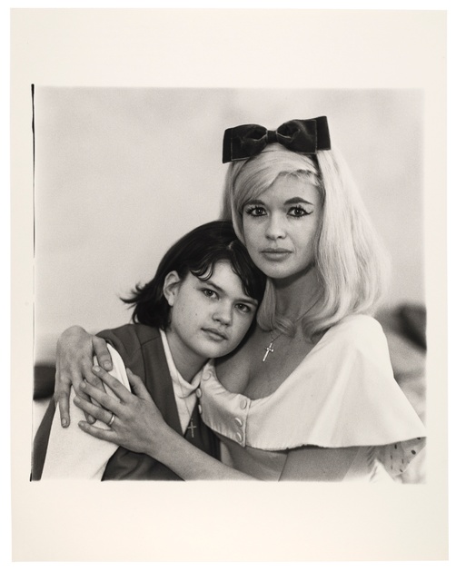 In a black and white photograph, a blond woman wearing a bow headband and winged eyeliner pulls a young, dark-haired girl into an embrace while both directly look at us.