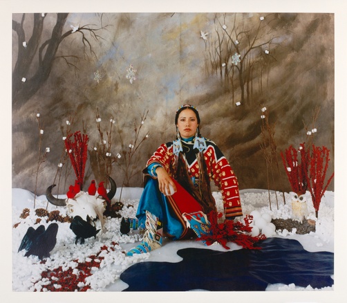 A young, tanned, Native woman sits in a fake snowy landscape wearing a bright red and blue dress with her hair in two braids.