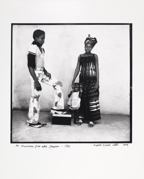 A black and white photograph of a young, Black family, a man, a woman, and a child, posed in front of a white wall.