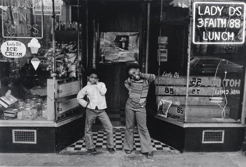 Two Black girls pose with their hands on their hips in front of an ice cream shop with an illuminated sign that reads “Lady D’s 3 Faith 88 Lunch.”