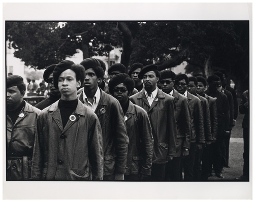 A black and white photograph of a group of Black men marching in line wearing similar attire.
