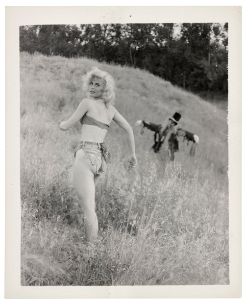 A black and white photograph of a white, young female running through a field looking over her shoulder with a scarecrow in the background.