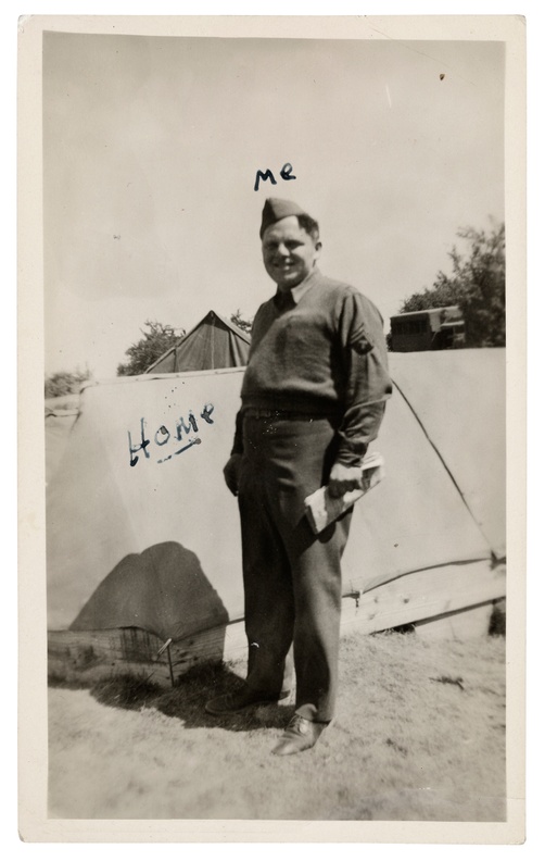 A grainy old black-and-white photograph shows a smiling white male in a military-looking uniform standing in front of a tent with “ME” handwritten above his head and “HOME” written on the tent.