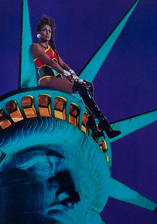 Against a blue background, a Black woman wearing a superhero costume and knee-high, heeled boots, sits on the crown of the Statue of Liberty as they both look into the distance.
