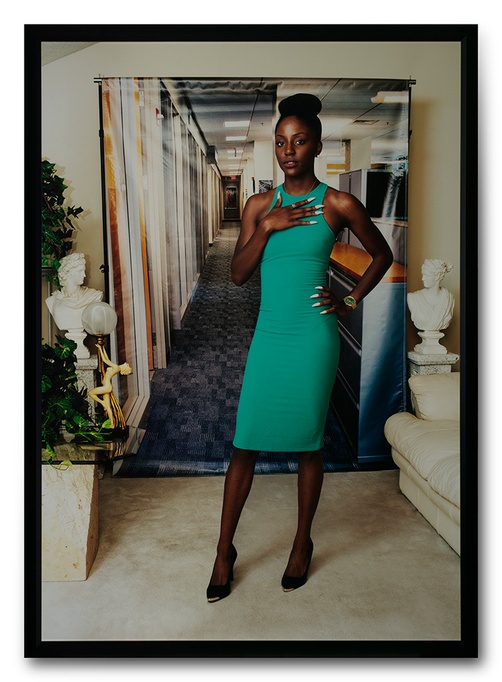In a living room-like space, a Black woman in a green dress stands looking out at the viewer in front of a photographic backdrop of an office setting. 