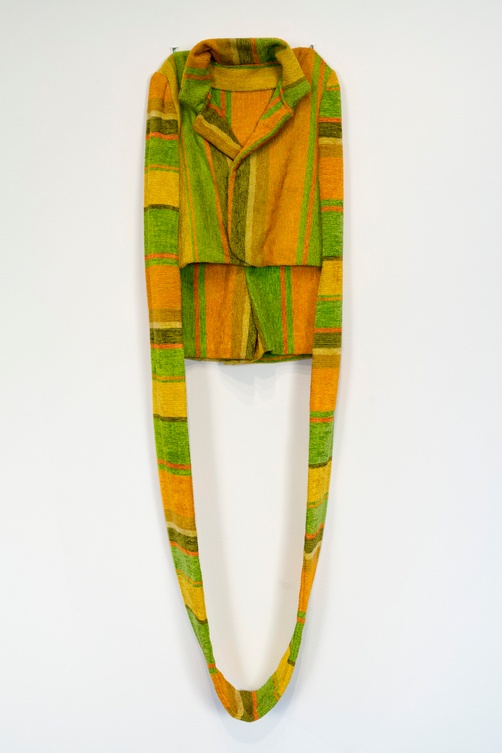 An orange, green, and yellow fabric artwork shaped as a tote bag hangs against a white background. The front of the bag resembles a collared shirt with the handle extending downward into a circular shape.