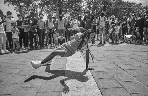 A black and white photograph of a young, black girl dancing surrounded by a circle of people looking on and cheering.