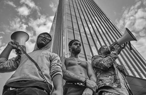 A black and white photograph of three, young, dark-skinned men standing side by side in front of a skyscraper. The two men on the ends hold megaphones.