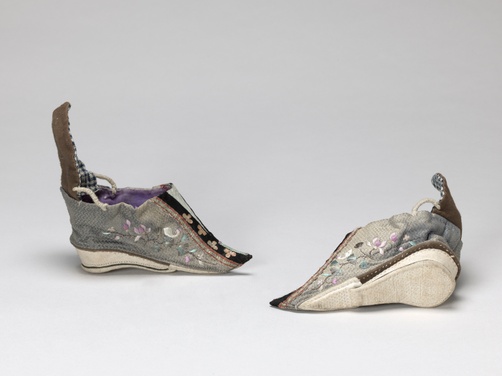 A gray pair of shoes with embroidered white birds, pastel purple lotus flowers, and pastel blue fanning leaves. Gold flower sequins on black fabric decorate the fronts.