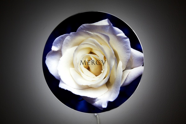 A white flower with purple edges is illuminated on a black, circular background, with “MERCY” appearing on the flower’s center. Light emanates from behind the sculpture on a grey wall.