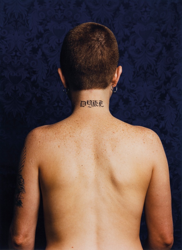 A nude, White, individual with a shaved head, hoop earrings, and a neck tattoo that says “Dyke,” stands with their back to us against a blue background. 