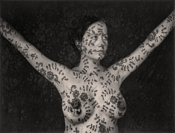 A black and white photo, covered in a pattern of flowers and foliage, of a woman with a nude upper body posing with arms up against a dark background.