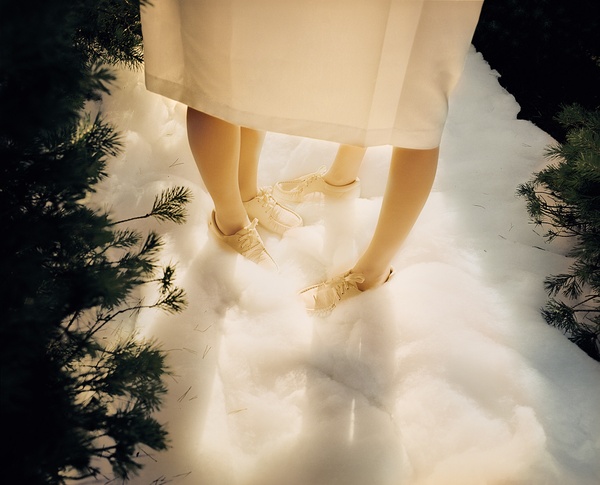 Two pairs of legs stick out from under a single skirt. Fake white snow covers the ground and green pine trees enter on both sides of the frame.
