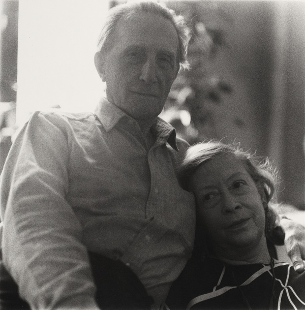 In a grainy black and white photograph, a woman rests her head on a man’s chest as he wraps his arms around her and looks directly toward us.