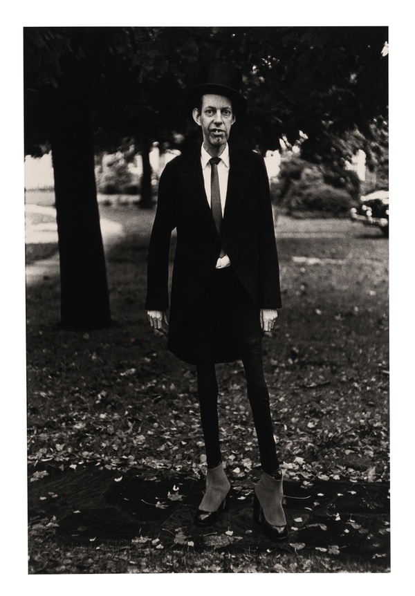 In a black and white photograph, a tall, slender man with a toothy grin stands in a park setting wearing a dark suit, heeled shoes, and a top hat.