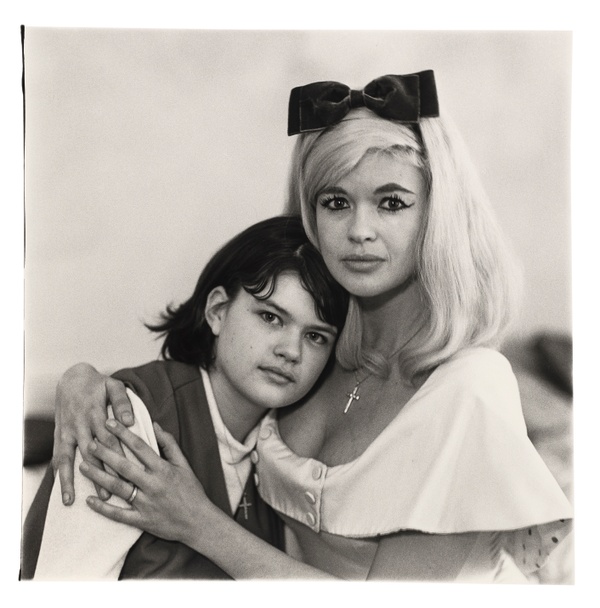 In a black and white photograph, a blond woman wearing a bow headband and winged eyeliner pulls a young, dark-haired girl into an embrace while both directly look at us.
