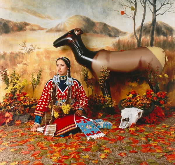 A young, tanned, Native woman sits in a fake fall landscape wearing a bright red and blue dress with her hair in two braids with a blow-up deer behind her.