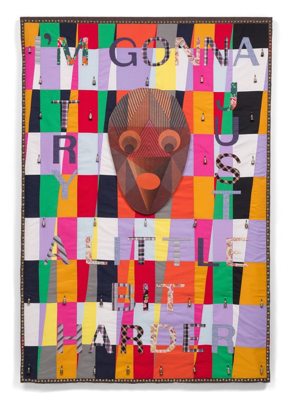 A colorful patchwork quilt with a brown, fabric mask in the center and cloth text "I'M GONNA TRY JUST A LITTLE BIT HARDER".