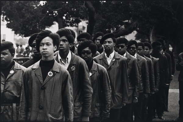 A black and white photograph of a group of Black men marching in line wearing similar attire.