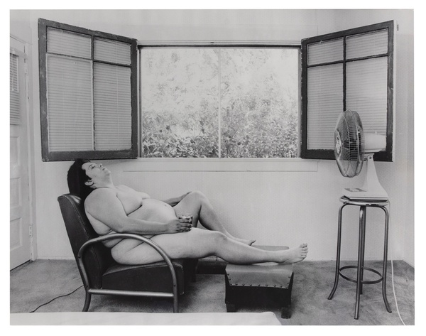 A black and white photograph of a nude, larger bodied woman reclining on a leather chair and ottoman in front of an open window with a curtain, as light fills the room. A fan is on a small table across from her.
