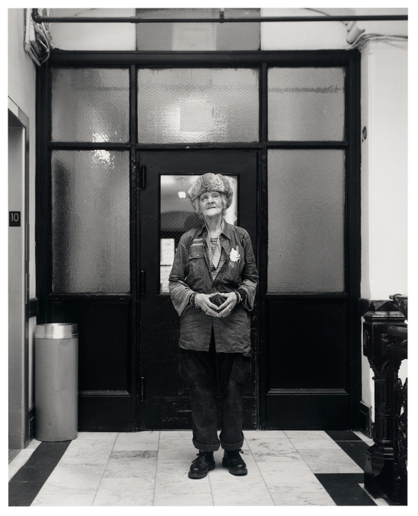 A black and white photo of someone standing in a marble-floored entryway. Windowpanes and a doorway frame the person, who wears a fur hat and faces the camera.